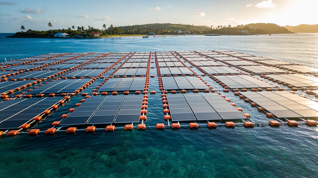 Aerial view of a large grid of floating solar panels in a calm blue tropical bay with anchored orange floats.